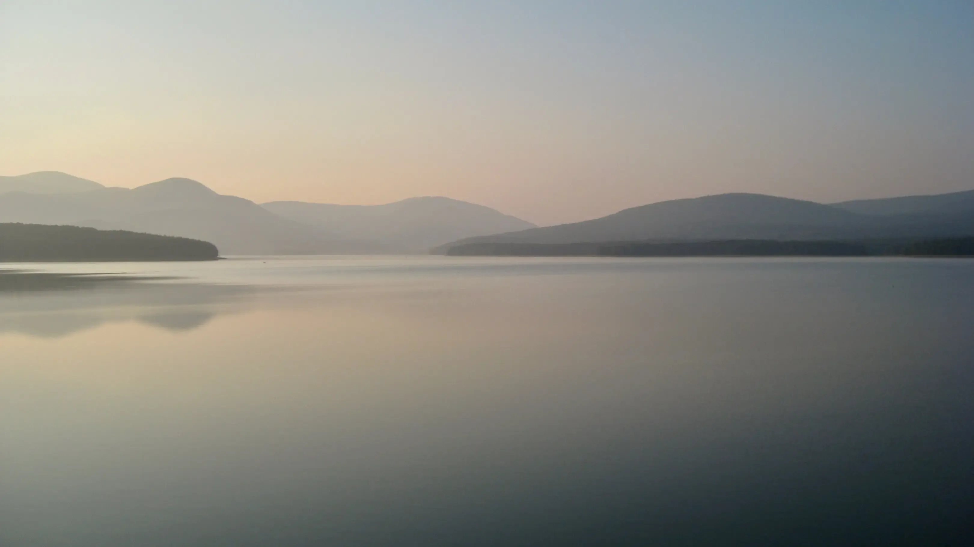 A tranquil body of water reflecting the sky, surrounded by natural scenery such as trees, mountains